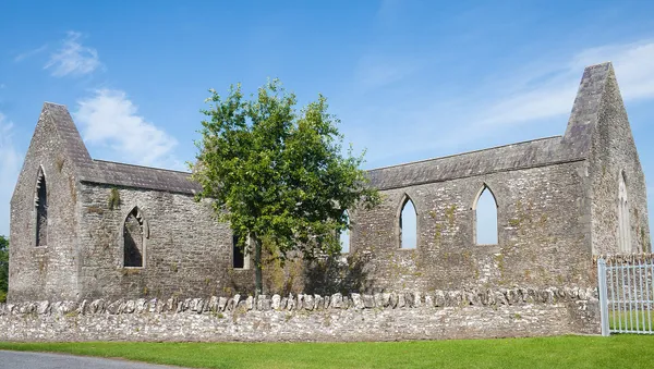 A roofless stone church with pointed window openings and a tree in front, under a blue sky.
