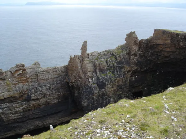 Jagged rock pinnacles and sheer cliffs above a calm sea, with a grassy slope in front.