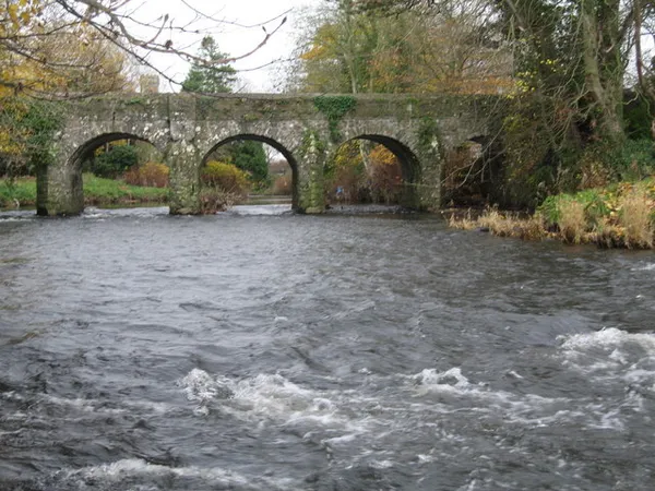 An ivy-covered stone bridge with arches over a fast-flowing river lined with trees.