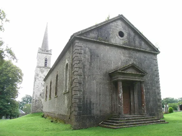 A grey stone church with a columned porch on a lawn, with a tall spired tower behind.