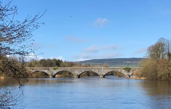 A long multi-arched stone bridge crossing a wide river, with trees and hills beyond under a blue sky.