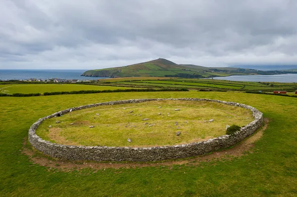 Eine ringförmige Steinmauer, die Gras und niedrige Steine auf einem Feld umschließt, mit Hügeln und dem Meer dahinter.