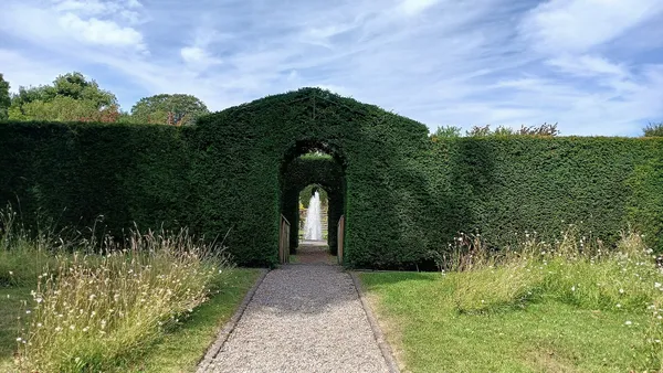 A gravel path leading through a tall clipped hedge arch towards a white fountain.