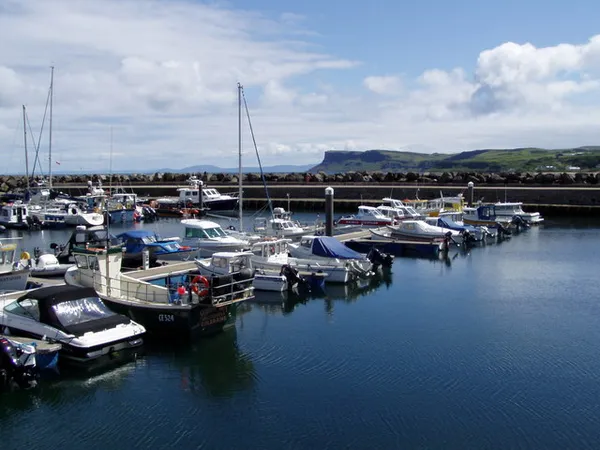 Small boats and yachts moored in a marina, with a headland in the distance under a partly cloudy sky.