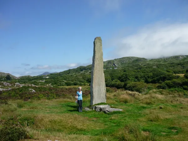 A tall standing stone in a grassy field with a person beside it and green hills behind.