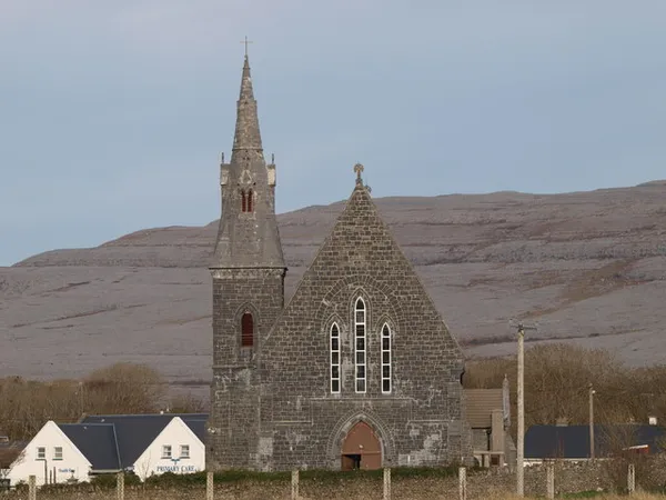 Une église en pierre grise avec une flèche haute devant un versant de calcaire nu et en terrasses.