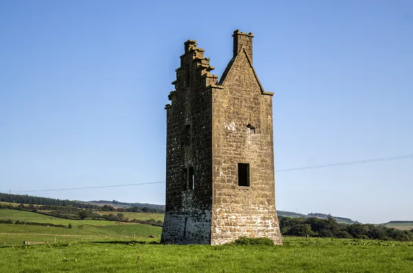A tall stone tower with stepped battlements standing in a green field under a clear blue sky.