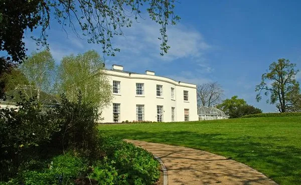 A white Georgian house with a conservatory above a sloping lawn and curving path, with trees around it.