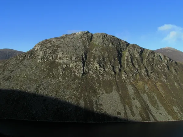 Una montaña escarpada y rocosa que se eleva sobre aguas oscuras bajo un cielo azul, con sus laderas inferiores en sombra.
