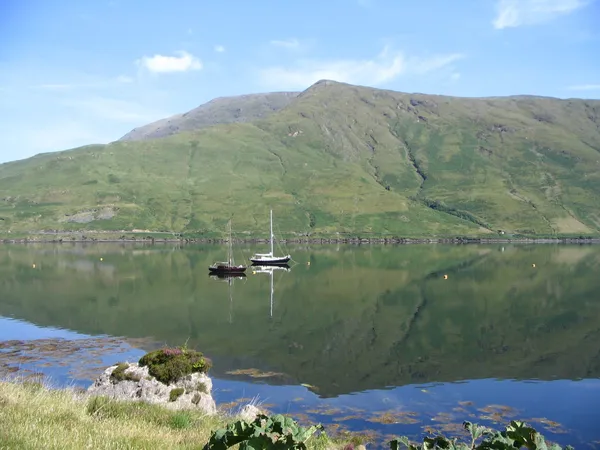 Una montagna verde riflessa in acqua calma, con due barche a vela ormeggiate e una riva rocciosa davanti.