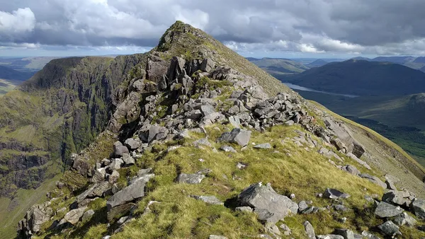 A narrow, rocky mountain ridge with grassy slopes rising to a peak, with steep cliffs and valleys beyond.