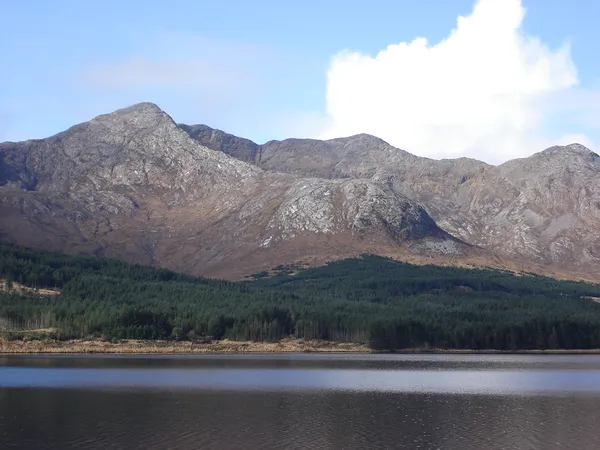 Rocky mountain peaks rising above a forested shore and a lake, with a white cloud in a blue sky.