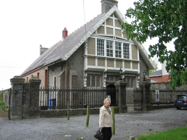 A stone hall with a steep slate roof and timbered gable behind railings, with a woman standing on the gravel in front.