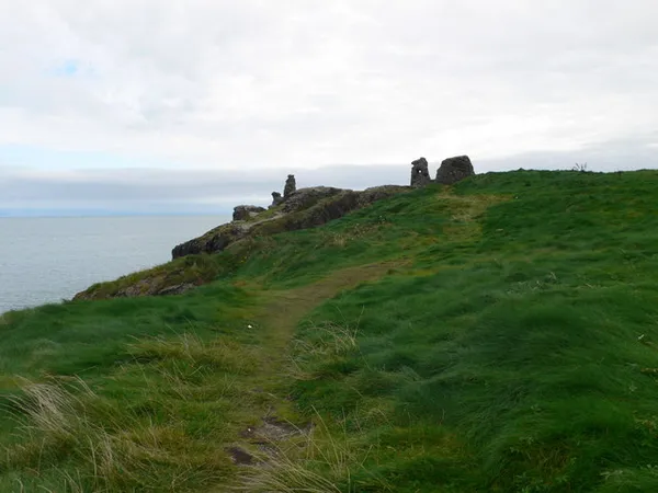 Un sentier herbeux menant à un promontoire vert jusqu'à de petites ruines en pierre au-dessus de la mer sous un ciel pâle.