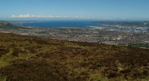 Brown heather slopes in the foreground looking down over a city and a wide blue sea lough.