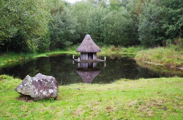Una piccola capanna con tetto di paglia su palafitte in uno stagno tranquillo, con un masso coperto di licheni sull'erba e alberi dietro.