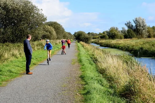 A paved path between grass verges beside a reedy canal, with people walking and cycling ahead.