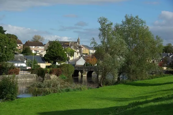 A stone bridge over a river in front of village houses and a church spire, seen from a grassy bank.