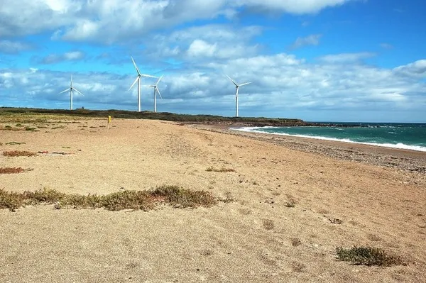 A sandy beach curving along the coast towards a low headland with wind turbines under a blue sky with white clouds.