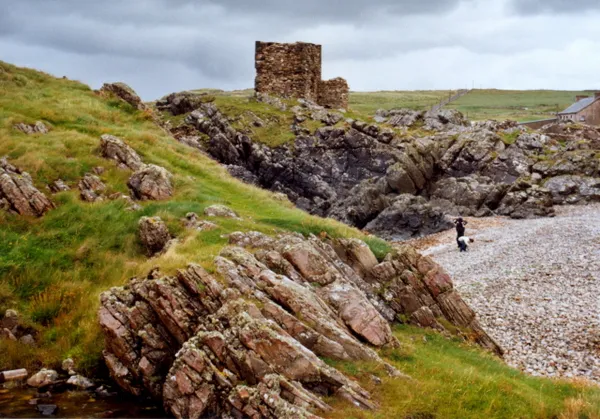 A ruined stone castle on a grassy headland above rugged rocks and a pebble beach.