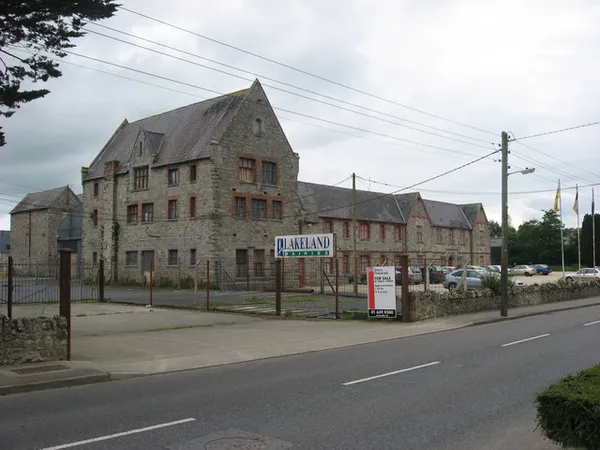 A large grey stone building with steep gables behind a railing, with a road in the foreground.