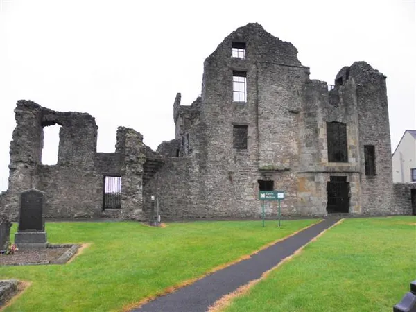 Roofless stone castle ruins with tall gables and empty windows behind a green lawn and a paved path.