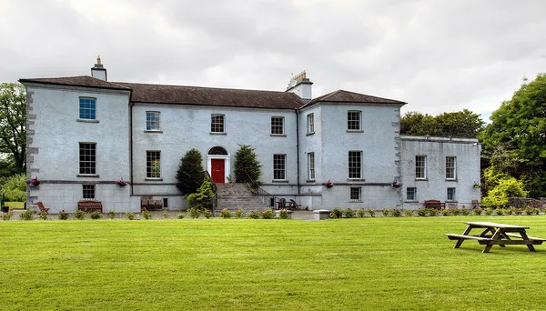 A long pale two-storey house with a red front door behind a wide green lawn with a picnic table.