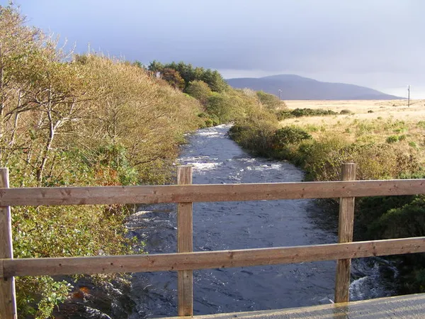 Un río poco profundo que fluye entre orillas cubiertas de hierba hacia una colina lejana, visto sobre una valla de madera.