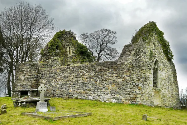 An ivy-topped ruined stone church in a grassy graveyard with a stone cross in front.
