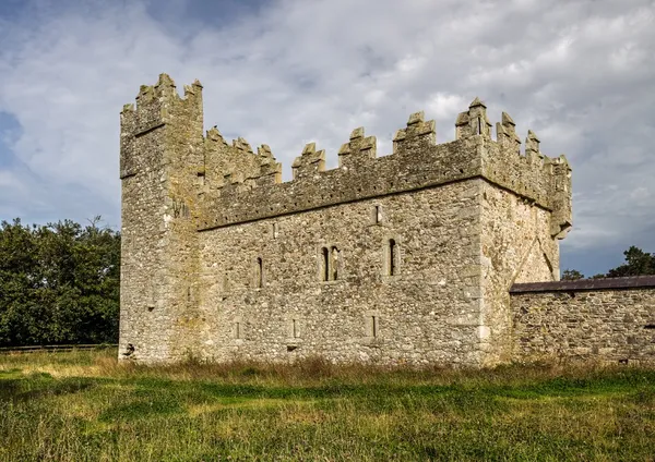 A square grey stone fortified house with crenellated battlements standing on a grassy field under a cloudy sky.