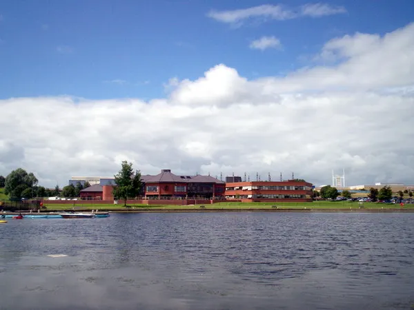 A wide lake in the foreground with low red-brick buildings on the far shore under a blue sky with white clouds.