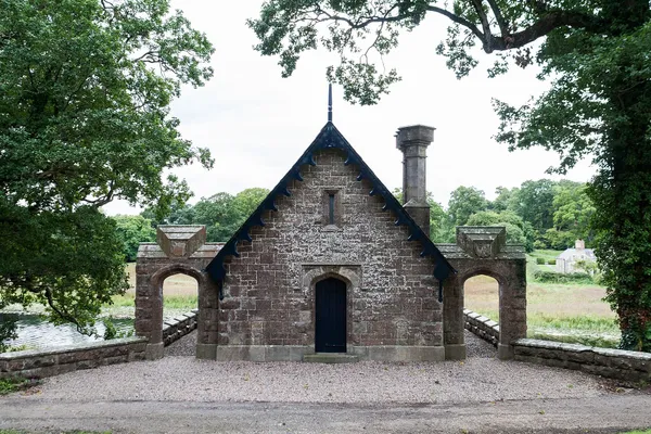 A stone boat house with a steep gable, tall chimney and arched side openings framed by trees.