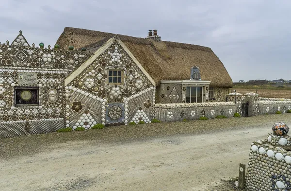 A thatched cottage and garden walls covered in patterns of seashells beside a gravel lane.
