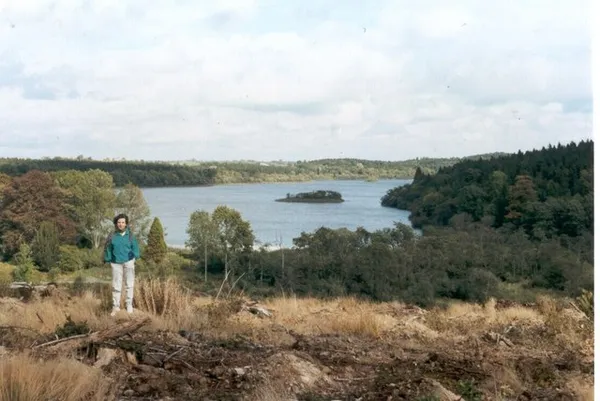 A walker stands on a cleared hillside above a lake with a small wooded island and forest beyond.