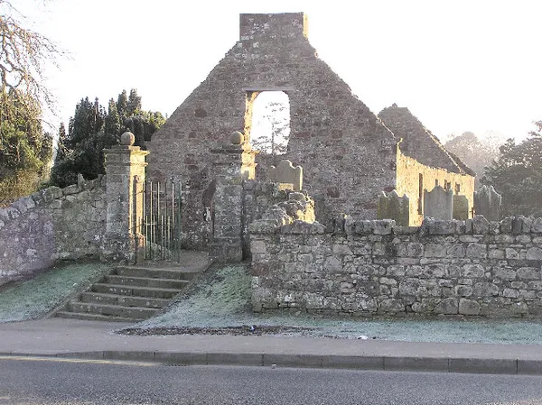 La ruina de piedra sin techo y con hastial de una iglesia detrás de un muro de cementerio con escalones y una puerta.