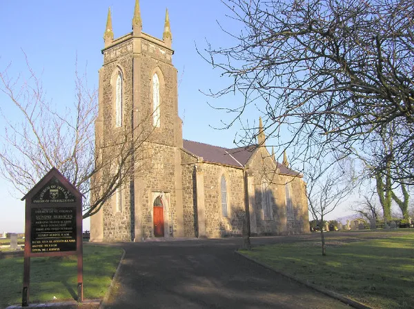 A stone church with a tall pinnacled tower and red door beside bare trees, with a noticeboard in front.
