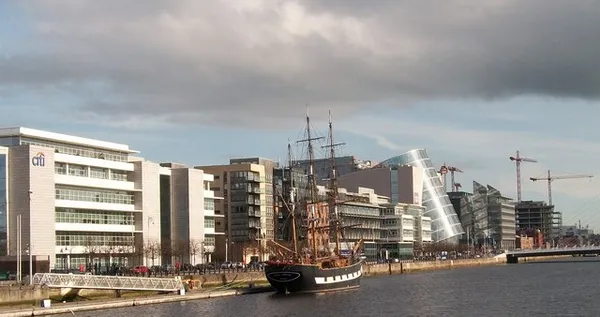 Un grand voilier amarré sur une rivière devant des immeubles de bureaux modernes et des grues de construction.