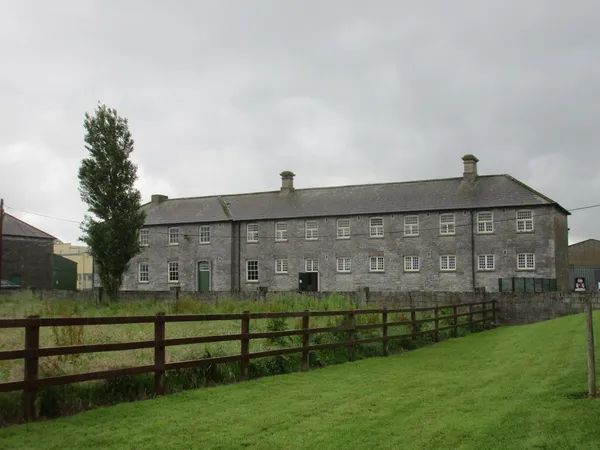 A long two-storey grey stone building with many windows beyond a wooden fence and grass.