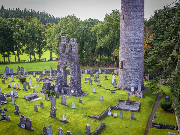 A tall stone round tower beside a ruined church gable in a graveyard of headstones and green grass.