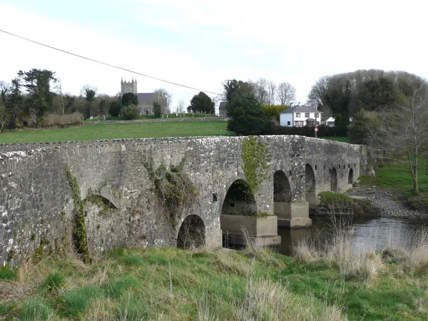 A multi-arched stone bridge over a river, with a church tower among trees on the bank behind.