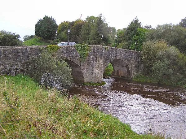 Un vieux pont de pierre en arche enjambant une rivière brune et rapide entre des rives herbeuses et arborées.