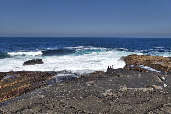 Ripiani rocciosi piatti e stratificati al bordo del mare con onde bianche che si infrangono e alcune persone in piedi sulle rocce.
