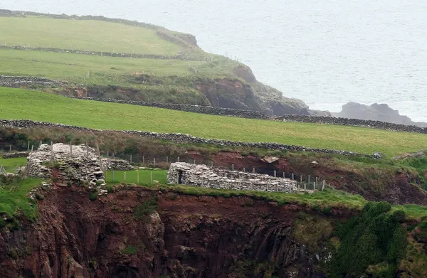 Niedrige Steinruinen auf einer grünen Klippe über erodierten roten Klippen, mit Feldern und einer Landzunge dahinter.