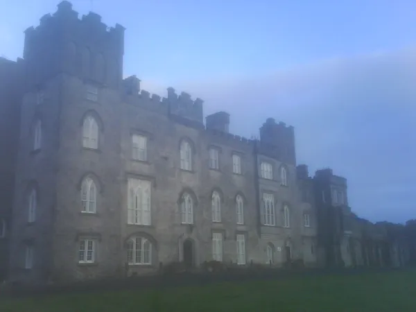 A long grey stone castle with crenellated towers and tall windows behind a lawn, softened by mist.