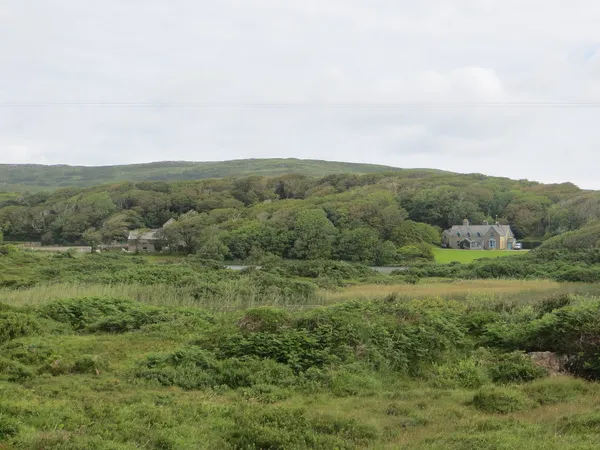 A grey stone house sits among dense trees in a green valley, rough grassland in the foreground.