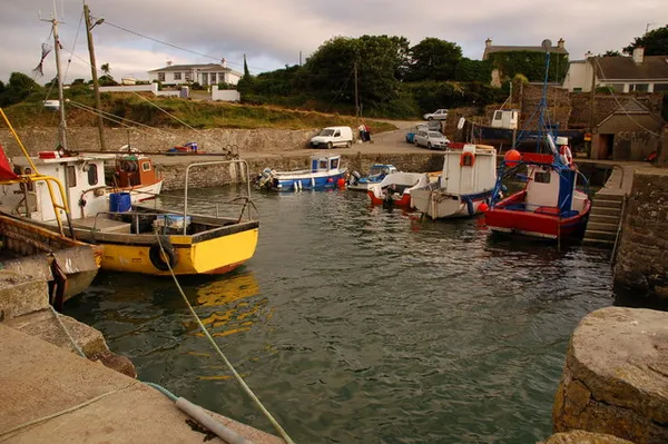 Small fishing boats moored in a stone-walled harbour with houses and trees behind.