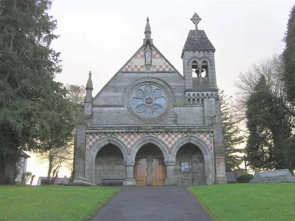 Stone Gothic church with a rose window and bell turret, set on a lawn among trees.