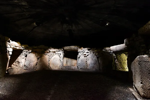 L'intérieur d'un tombeau à couloir avec des murs en pierre et un toit en dôme bas éclairé d'en haut.