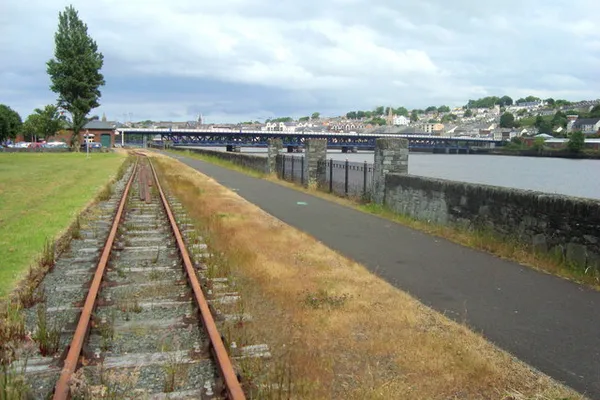 A disused railway track beside a paved path and river wall, with a bridge and hillside houses beyond.