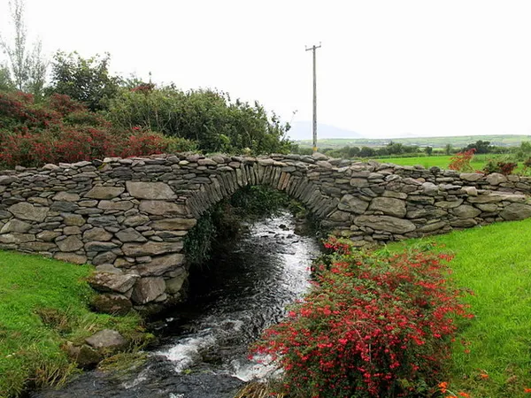 Eine niedrige Trockensteinbogenbrücke über einen kleinen Bach, davor rote Blumen und grünes Gras.